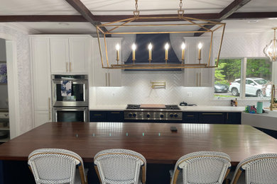 This is an example of a large modern l-shaped kitchen in Baltimore with a belfast sink, shaker cabinets, white cabinets, engineered stone countertops, white splashback, metro tiled splashback, integrated appliances, medium hardwood flooring, an island, brown floors, white worktops and exposed beams.