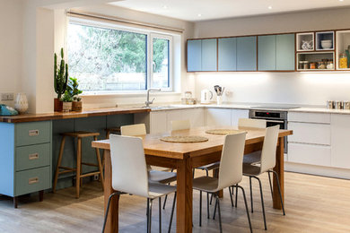 This is an example of a midcentury l-shaped eat-in kitchen in Hampshire with flat-panel cabinets, blue cabinets, wood benchtops, white splashback, stainless steel appliances, light hardwood floors and no island.