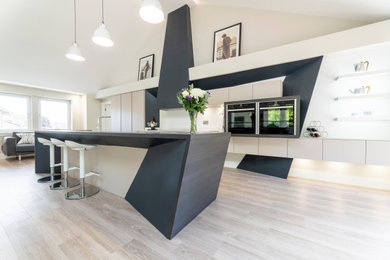 Design ideas for a large contemporary galley open plan kitchen in Edinburgh with a built-in sink, flat-panel cabinets, white cabinets, quartz worktops, white splashback, stone slab splashback, black appliances, light hardwood flooring, an island, beige floors and white worktops.