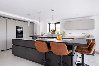 This is an example of a large contemporary l-shaped open plan kitchen in Gloucestershire with an integrated sink, flat-panel cabinets, grey cabinets, composite countertops, grey splashback, engineered quartz splashback, black appliances, porcelain flooring, an island and grey worktops.