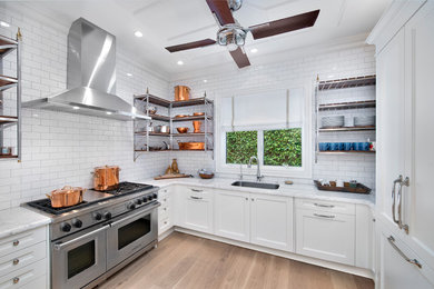 This is an example of a beach style u-shaped kitchen in Cleveland with an undermount sink, shaker cabinets, white cabinets, white splashback, subway tile splashback, stainless steel appliances, light hardwood floors and no island.