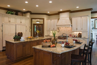 Photo of a large traditional l-shaped separate kitchen in St Louis with a double-bowl sink, raised-panel cabinets, beige cabinets, granite benchtops, beige splashback, stone tile splashback, panelled appliances, dark hardwood floors, multiple islands and brown floor.