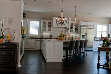 This is an example of a large traditional l-shaped eat-in kitchen in Orange County with a drop-in sink, white cabinets, granite benchtops, stainless steel appliances, with island, raised-panel cabinets, white splashback, mosaic tile splashback, dark hardwood floors, brown floor and black benchtop.