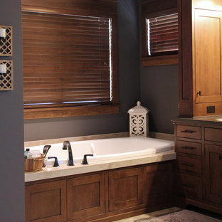 Modelo de cuarto de baño principal, de estilo americano, grande, con lavabo bajoencimera, armarios con paneles empotrados, puertas de armario de madera oscura, encimera de acrílico, bañera encastrada, ducha abierta, sanitario de dos piezas, baldosas y/o azulejos de porcelana, paredes grises y suelo de baldosas de porcelana