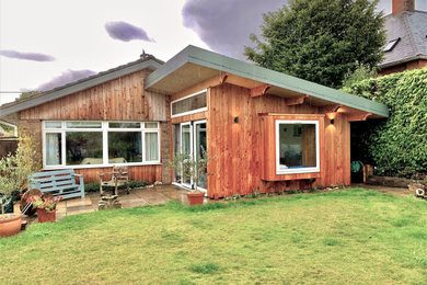 Medium sized and brown scandinavian bungalow rear house exterior in Wiltshire with wood cladding, a half-hip roof and a grey roof.