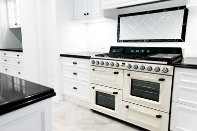 Design ideas for a large traditional galley open plan kitchen in Brisbane with a farmhouse sink, shaker cabinets, white cabinets, solid surface benchtops, white splashback, subway tile splashback, white appliances, light hardwood floors, with island, grey floor and black benchtop.
