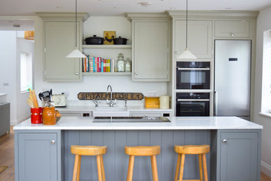 This is an example of a large country single-wall kitchen in London with a farmhouse sink, shaker cabinets, marble benchtops, white splashback, marble splashback, stainless steel appliances, light hardwood floors, with island, white benchtop and grey cabinets.