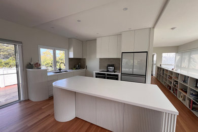 Large modern kitchen pantry in Sydney with an undermount sink, flat-panel cabinets, white cabinets, solid surface benchtops, grey splashback, stainless steel appliances, light hardwood floors, with island, brown floor and white benchtop.