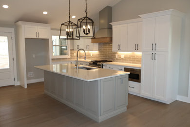 Example of a large minimalist l-shaped light wood floor, brown floor and vaulted ceiling open concept kitchen design in Other with an undermount sink, shaker cabinets, white cabinets, quartzite countertops, beige backsplash, ceramic backsplash, stainless steel appliances, an island and white countertops