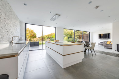 Design ideas for a large contemporary grey and white l-shaped open plan kitchen in Essex with a built-in sink, flat-panel cabinets, white cabinets, quartz worktops, grey splashback, stone tiled splashback, black appliances, porcelain flooring, an island, grey floors, white worktops and a feature wall.