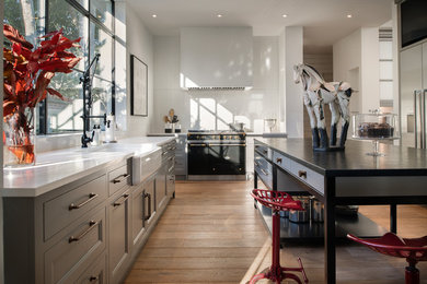 Open concept kitchen - mid-sized cottage l-shaped medium tone wood floor and beige floor open concept kitchen idea in Orlando with a farmhouse sink, recessed-panel cabinets, gray cabinets, solid surface countertops, window backsplash, black appliances and an island