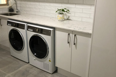 Design ideas for a mid-sized contemporary galley dedicated laundry room in Other with a single-bowl sink, flat-panel cabinets, white cabinets, solid surface benchtops, white splashback, ceramic splashback, white walls, porcelain floors, a side-by-side washer and dryer, grey floor and white benchtop.