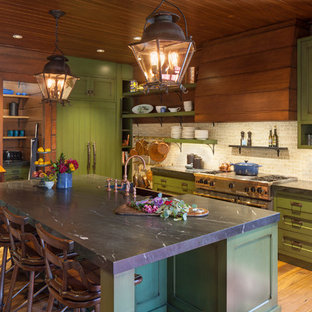 Modelo de cocina rústica, extra grande, con fregadero sobremueble, armarios estilo shaker, puertas de armario verdes, salpicadero beige, electrodomésticos de acero inoxidable, suelo de madera en tonos medios, una isla y encimeras negras