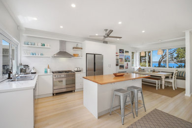 Photo of a beach style l-shaped kitchen in Central Coast with a drop-in sink, flat-panel cabinets, white cabinets, white splashback, subway tile splashback, stainless steel appliances, light hardwood floors, with island, beige floor and white benchtop.