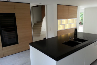 Exemple d'une grande cuisine américaine tendance avec un évier intégré, un placard à porte affleurante, des portes de placard blanches, un plan de travail en granite, une crédence blanche, une crédence en feuille de verre, un électroménager noir, un sol en carrelage de céramique, îlot, un sol gris et plan de travail noir.