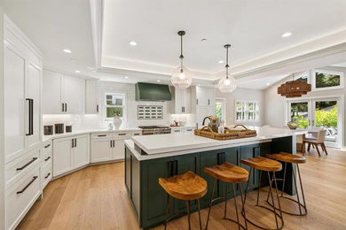 Cottage l-shaped medium tone wood floor, brown floor and tray ceiling eat-in kitchen photo in San Francisco with a farmhouse sink, shaker cabinets, white cabinets, quartz countertops, white backsplash, ceramic backsplash, paneled appliances, an island and white countertops