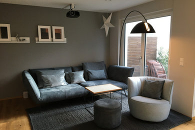 Imagen de sala de estar contemporánea grande con paredes grises, suelo de madera clara, televisor colgado en la pared y suelo marrón