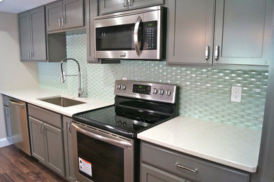 Example of a mid-sized minimalist galley medium tone wood floor kitchen pantry design in Cleveland with a single-bowl sink, shaker cabinets, gray cabinets, quartz countertops, green backsplash, glass sheet backsplash, stainless steel appliances and an island