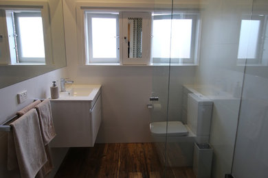 Photo of a mid-sized modern master bathroom in Sydney with flat-panel cabinets, white cabinets, solid surface benchtops, brown tile, porcelain tile, a corner shower, a one-piece toilet, a wall-mount sink, white walls and porcelain floors.