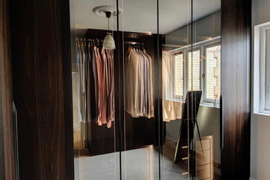 Inspiration for a large modern gender neutral dressing room in London with glass-front cabinets, dark wood cabinets, carpet, grey floors and feature lighting.