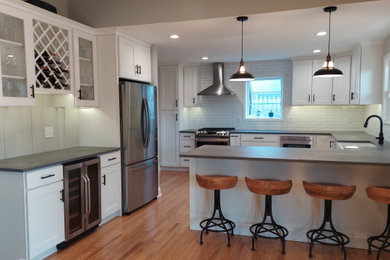 Photo of a large farmhouse u-shaped kitchen/diner in Philadelphia with a belfast sink, shaker cabinets, white cabinets, engineered stone countertops, white splashback, metro tiled splashback, stainless steel appliances, light hardwood flooring, a breakfast bar, brown floors, grey worktops and a vaulted ceiling.