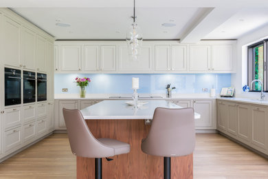 Photo of a large modern grey and brown u-shaped open plan kitchen in Surrey with a submerged sink, shaker cabinets, grey cabinets, quartz worktops, blue splashback, glass sheet splashback, black appliances, vinyl flooring, an island, brown floors, white worktops, exposed beams and a feature wall.