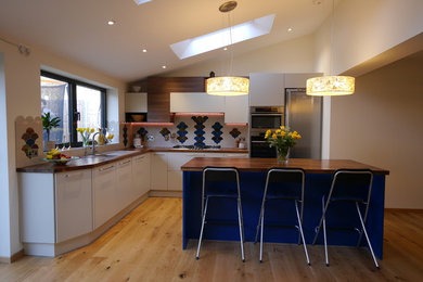 ロンドンにある低価格の広いモダンスタイルのおしゃれなキッチン (ドロップインシンク、フラットパネル扉のキャビネット、青いキャビネット、木材カウンター、青いキッチンパネル、セラミックタイルのキッチンパネル、シルバーの調理設備、淡色無垢フローリング、ベージュの床、茶色いキッチンカウンター) の写真