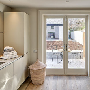 Exempel på en stor modern linjär tvättstuga, med en enkel diskho, släta luckor, vita skåp, vita väggar, mellanmörkt trägolv och en tvättmaskin och torktumlare bredvid varandra