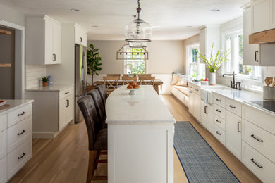 Inspiration for a traditional kitchen/diner in Boston with a belfast sink, shaker cabinets, white cabinets, engineered stone countertops, white splashback, metro tiled splashback, stainless steel appliances, medium hardwood flooring, an island and white worktops.