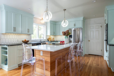 This is an example of a mid-sized transitional u-shaped kitchen in New York with a farmhouse sink, shaker cabinets, soapstone benchtops, white splashback, ceramic splashback, medium hardwood floors, with island, brown floor, black benchtop, turquoise cabinets and stainless steel appliances.