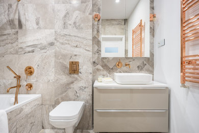 Small contemporary grey and cream ensuite bathroom in London with flat-panel cabinets, beige cabinets, a built-in bath, a wall mounted toilet, brown tiles, stone tiles, multi-coloured walls, ceramic flooring, a vessel sink, brown floors, a feature wall, a single sink, a floating vanity unit and a drop ceiling.