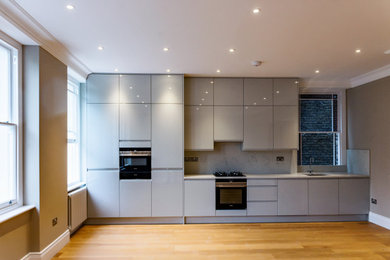 Photo of a contemporary grey and white kitchen in London with a single-bowl sink, granite splashback, black appliances, medium hardwood flooring, brown floors and feature lighting.