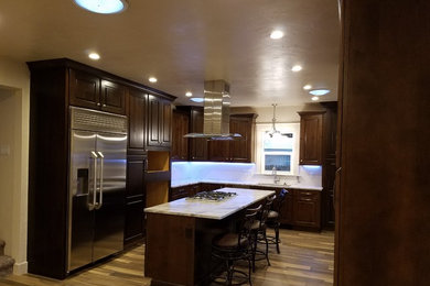 Elegant light wood floor kitchen photo in Denver with raised-panel cabinets, dark wood cabinets, white backsplash, stainless steel appliances and an island