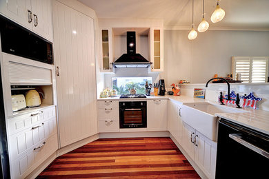 Small midcentury u-shaped separate kitchen in Brisbane with a farmhouse sink, glass-front cabinets, white cabinets, quartz benchtops, window splashback, black appliances, dark hardwood floors and white benchtop.