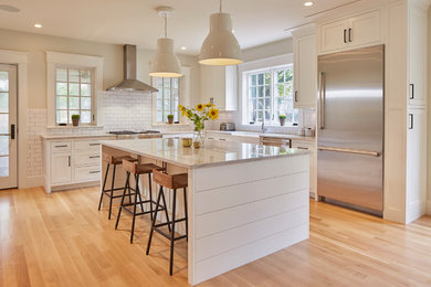 Inspiration for a transitional l-shaped medium tone wood floor and brown floor kitchen remodel in Boston with a farmhouse sink, shaker cabinets, white cabinets, white backsplash, subway tile backsplash, stainless steel appliances, an island and white countertops