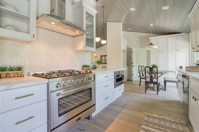 Photo of a mid-sized country galley eat-in kitchen in San Francisco with an undermount sink, shaker cabinets, white cabinets, quartz benchtops, white splashback, ceramic splashback, stainless steel appliances, light hardwood floors, a peninsula, grey floor and white benchtop.