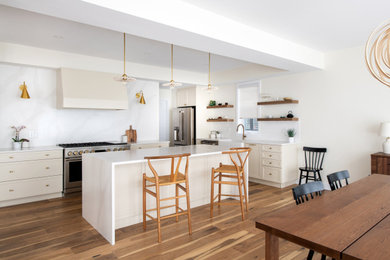 Large danish l-shaped medium tone wood floor and brown floor eat-in kitchen photo in Toronto with a farmhouse sink, shaker cabinets, white cabinets, quartz countertops, white backsplash, quartz backsplash, stainless steel appliances, an island and white countertops