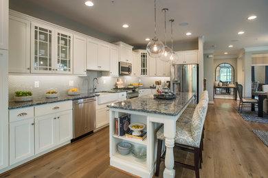 Mid-sized transitional l-shaped open plan kitchen in Philadelphia with a farmhouse sink, recessed-panel cabinets, white cabinets, quartz benchtops, grey splashback, stainless steel appliances, light hardwood floors and with island.