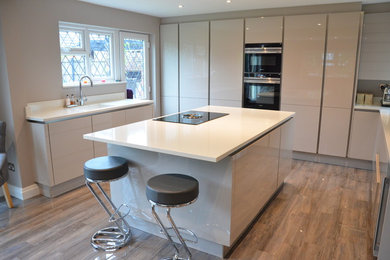 This is an example of a large contemporary u-shaped kitchen/diner in Essex with a submerged sink, flat-panel cabinets, beige cabinets, white splashback, black appliances, porcelain flooring, an island and beige floors.