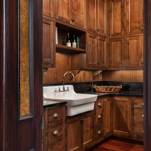 他の地域の大きいヴィクトリアン調のおしゃれな家事室 (エプロンフロントシンク、シェーカースタイル扉のキャビネット、濃色木目調キャビネット、人工大理石カウンター、茶色い壁、左右配置の洗濯機・乾燥機、濃色無垢フローリング、茶色い床) の写真