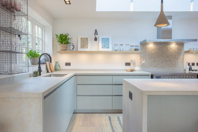 Inspiration for a modern u-shaped kitchen/diner in Hampshire with an integrated sink, flat-panel cabinets, green cabinets, engineered stone countertops, beige splashback, brick splashback, stainless steel appliances, medium hardwood flooring, an island, beige worktops and feature lighting.
