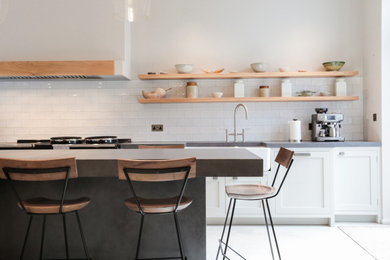 Inspiration for a medium sized modern kitchen/diner in London with a belfast sink, shaker cabinets, white cabinets, concrete worktops, white splashback, porcelain flooring, an island, grey worktops, a chimney breast and white appliances.