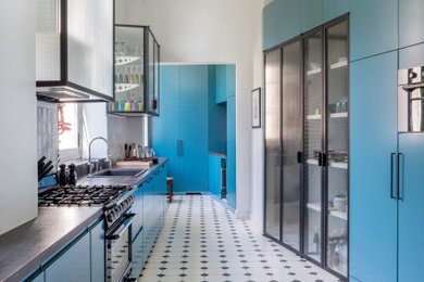 Exemple d'une cuisine parallèle tendance avec un placard à porte vitrée, des portes de placard bleues, une crédence grise, un électroménager noir, un plan de travail gris et fenêtre au-dessus de l'évier.