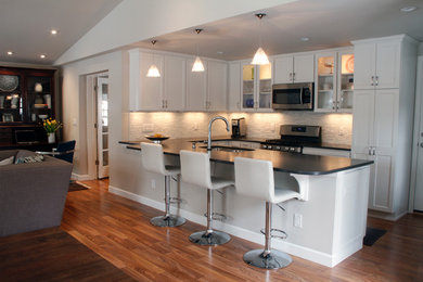 Example of a mid-sized trendy galley medium tone wood floor open concept kitchen design in Denver with a double-bowl sink, shaker cabinets, white cabinets, quartz countertops, white backsplash, glass tile backsplash, stainless steel appliances and a peninsula