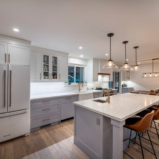 Mid-sized contemporary single-wall kitchen pantry in Toronto with a farmhouse sink, shaker cabinets, white cabinets, quartz benchtops, white splashback, brick splashback, stainless steel appliances, medium hardwood floors, with island, brown floor and white benchtop.
