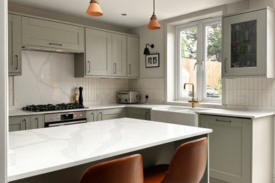 Inspiration for a medium sized contemporary l-shaped open plan kitchen in London with a belfast sink, shaker cabinets, green cabinets, composite countertops, white splashback, ceramic splashback, black appliances, porcelain flooring, an island, beige floors, white worktops and feature lighting.