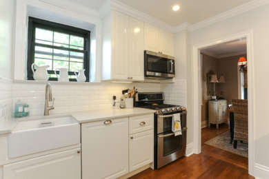 Photo of a medium sized classic single-wall open plan kitchen in New York with a belfast sink, raised-panel cabinets, white cabinets, composite countertops, white splashback, metro tiled splashback, stainless steel appliances, medium hardwood flooring, no island, brown floors and white worktops.