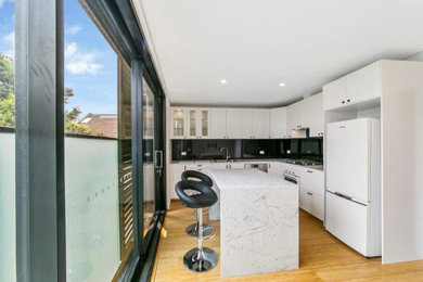 Inspiration for a mid-sized contemporary l-shaped eat-in kitchen in Sydney with a single-bowl sink, flat-panel cabinets, white cabinets, quartz benchtops, black splashback, glass tile splashback, stainless steel appliances, light hardwood floors, with island, brown floor and white benchtop.