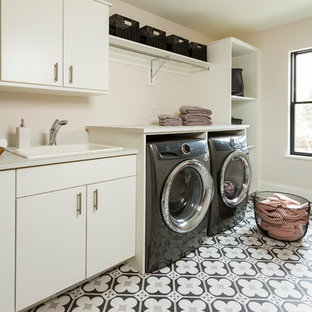 ミネアポリスの大きいトランジショナルスタイルのおしゃれな洗濯室 (ドロップインシンク、フラットパネル扉のキャビネット、白いキャビネット、ラミネートカウンター、ピンクの壁、左右配置の洗濯機・乾燥機、マルチカラーの床、コンクリートの床、白いキッチンカウンター) の写真