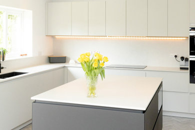 Medium sized contemporary u-shaped kitchen/diner in Manchester with a single-bowl sink, flat-panel cabinets, white cabinets, quartz worktops, white splashback, black appliances, light hardwood flooring, an island, grey floors and white worktops.